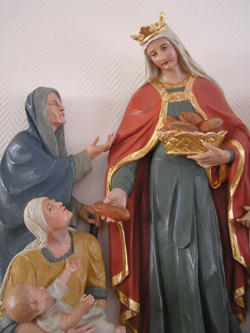 Eine Statur der Heiligen Elisabeth zeigt sie beim Austeilen von Brot. In der linken Hand h�lt sie einen Brotkorb. Ihre Linke reicht einem Bed�rftigen ein St�ck davon.  Vor den Beschenkten hockt eine ebenfalls bed�rftige Frau mit einem Kind auf dem Scho�.  Mutter und Kind strecken ihre Arme Elisabeth entgegen.