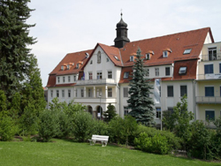 Hinter einer saftig gr�nen Wiese, begrenzt mit Streuchern und einem an der linksen Seite stehenden Baum, erscheint das Geb�ude des Hospizes. Es ist ab der 2. Ebene zu sehen. eine helle Fassade wird in der Mitte durch einen Vorbau mit Balkonen versch�nert. Ein Hohes rotes Ziegeldach �ber weitere 2 Stockwerke ist mit 4 Erknern versehen. �ber dem Vorbau ist ein heller Giebel errichtet.