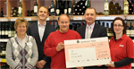 Vor einem Weinregal des REWE-Marktes haben sich die Anwesenden zu einem Gruppenfoto versammelt. Die Mitarbeiter des Marktes halten gemeinsam einen ?bergro?en Scheck. Der Spendenbetrag von 2000 ? ist gut erkennbar. Allen Beteiligten steht die Freude ?ber diese Spende ins Gesicht geschrieben. 