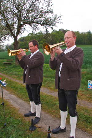 Wir sehen 2 Musikanten, wie sie auf ihren Trompeten spielen. Sie stehen auf einem Weg im Hopperain, der durch 2 graue Fahrspuren gekennzeichnet ist. Hinter den Musikanten ist ein Obstbaum zu sehen. Unter ihm l�dt eine Bank zum verweilen ein. Ein Waldrand hinter einem gr�n bestellten Feld schlie�t das Bild am Horizont ab.   